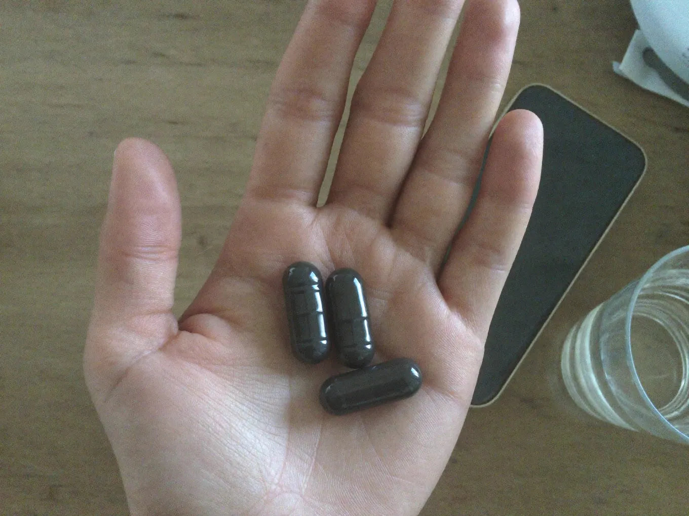 Hand holding three capsules next to a phone on a desk